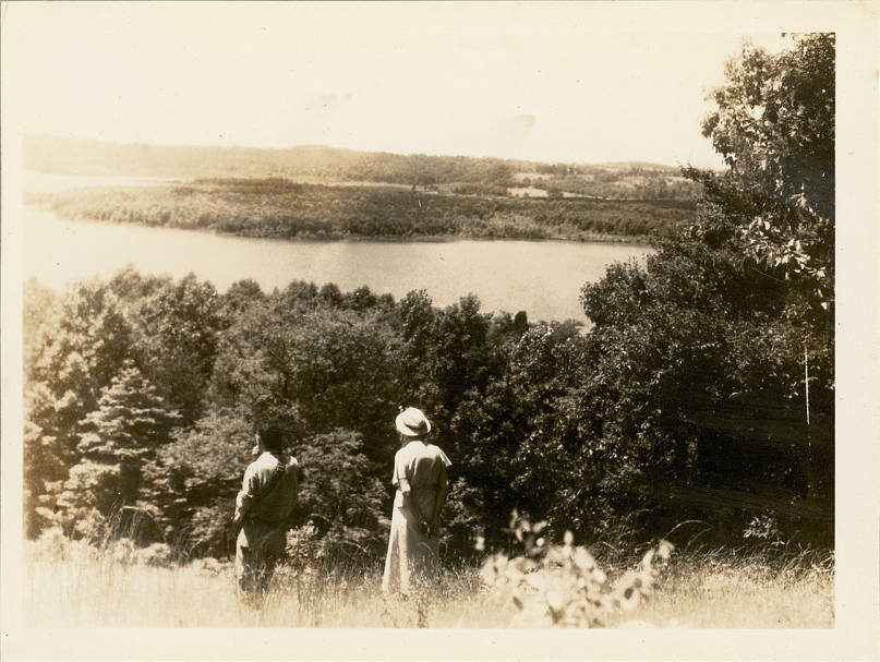 Photograph of a man and a woman standing on a hill overlooking Loch Raven Reservoir and the surrounding wooded landscape taken by Irving Edward Hampe on June 12, 1938, in Loch Raven, Maryland. From Photograph Record album BE, negative number Ec20. This photo can be seen with its original notes in the album under identifier nhsm0011.